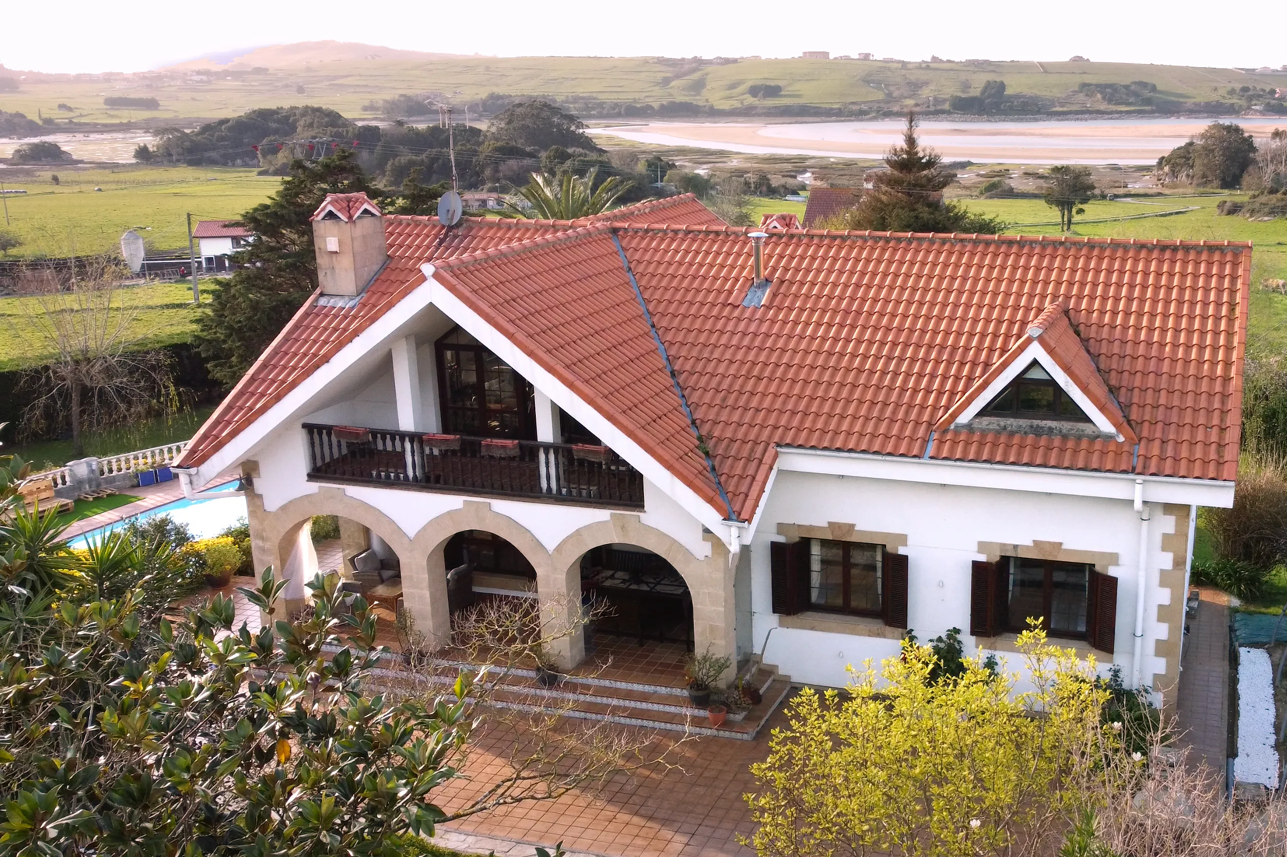 Vista exterior de una casa unifamiliar de dos plantas con tejado de teja roja, porche de arcos de piedra y terraza, situada frente a un paisaje verde con estuario al fondo.